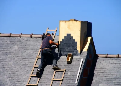 Couvreur sur une &eacute;chelle de toit posant des ardoises pr&egrave;s d'une chemin&eacute;e en pierre sous un ciel bleu