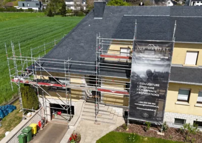 Vue a&eacute;rienne d'une maison individuelle &eacute;chafaud&eacute;e avec toiture en ardoise neuve et b&acirc;che Lux Habitat, en bordure de champ